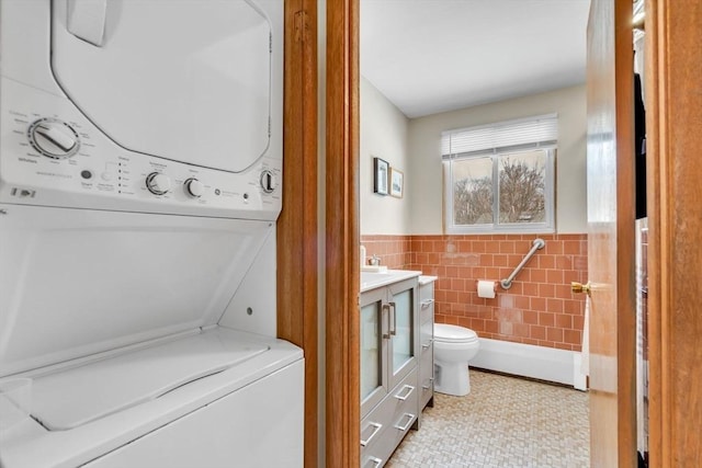 bathroom featuring stacked washer and clothes dryer, tile walls, toilet, wainscoting, and vanity