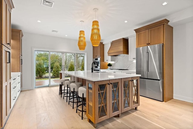 kitchen with brown cabinets, custom range hood, visible vents, appliances with stainless steel finishes, and a kitchen island with sink