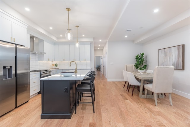 kitchen with pendant lighting, a center island with sink, wall chimney range hood, white cabinetry, and stainless steel appliances