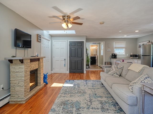 living room featuring a baseboard heating unit, light hardwood / wood-style flooring, ceiling fan, a fireplace, and a skylight