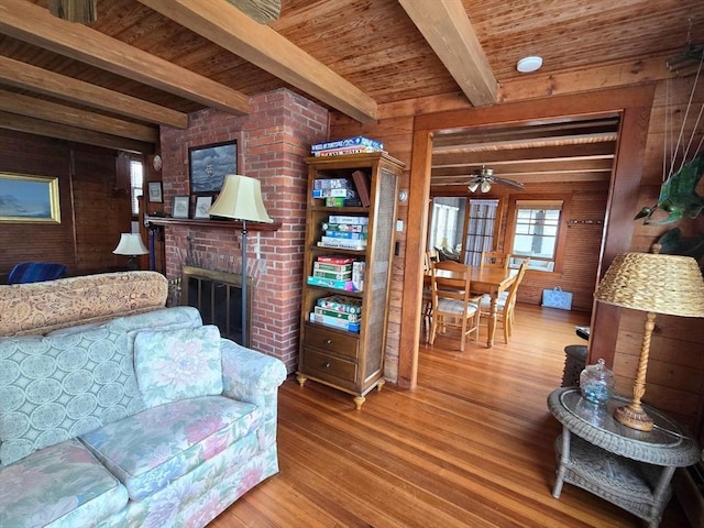 living room featuring wood walls, a brick fireplace, hardwood / wood-style flooring, ceiling fan, and beamed ceiling