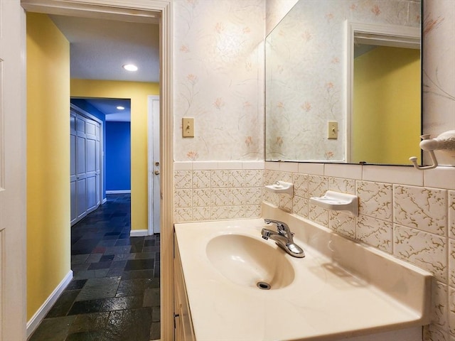 bathroom featuring wallpapered walls, stone tile floors, baseboards, vanity, and recessed lighting