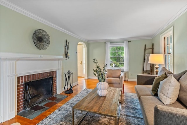 living room featuring crown molding, wood finished floors, arched walkways, a brick fireplace, and baseboards