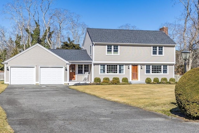 colonial-style house featuring aphalt driveway, roof with shingles, an attached garage, a front yard, and a chimney