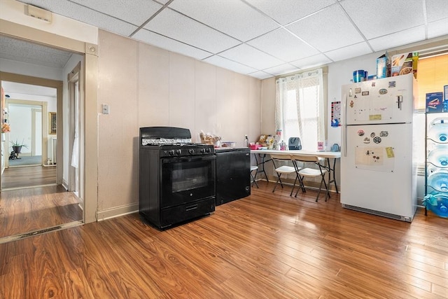 kitchen with black gas range oven, white refrigerator, a drop ceiling, and wood-type flooring