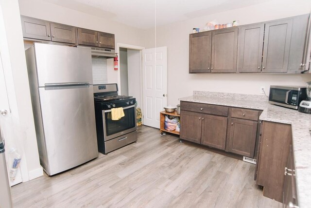 kitchen with appliances with stainless steel finishes, light stone countertops, dark brown cabinets, light wood finished floors, and under cabinet range hood