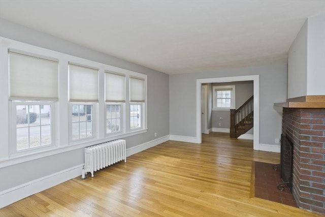 unfurnished living room featuring stairway, baseboards, light wood finished floors, radiator heating unit, and a brick fireplace