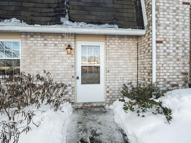 snow covered property entrance with roof with shingles and brick siding