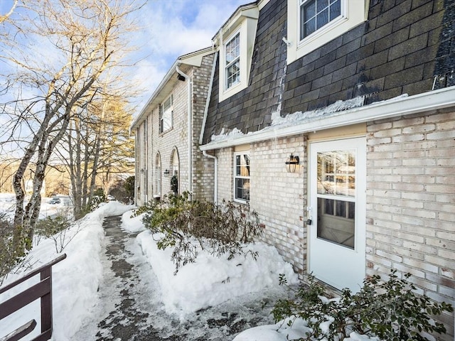 view of snow covered exterior with mansard roof, a shingled roof, and brick siding