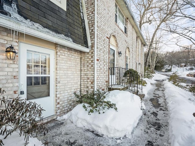 snow covered property featuring brick siding and roof with shingles