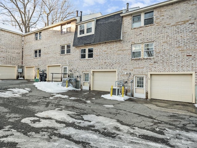 rear view of house featuring driveway, a garage, mansard roof, roof with shingles, and brick siding