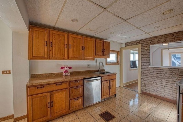 kitchen with sink, light tile patterned floors, brick wall, a drop ceiling, and stainless steel dishwasher