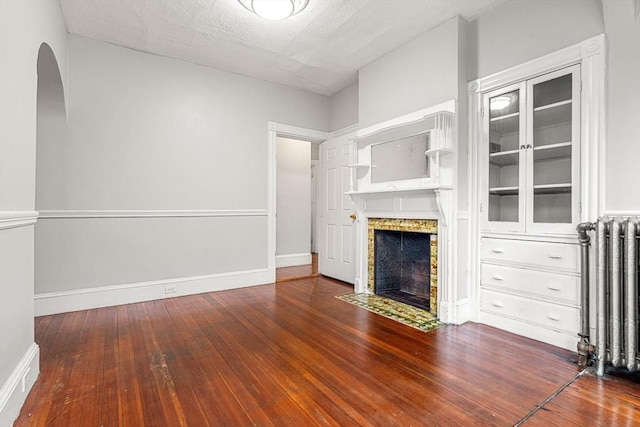 unfurnished living room featuring radiator, a fireplace with flush hearth, hardwood / wood-style flooring, arched walkways, and baseboards