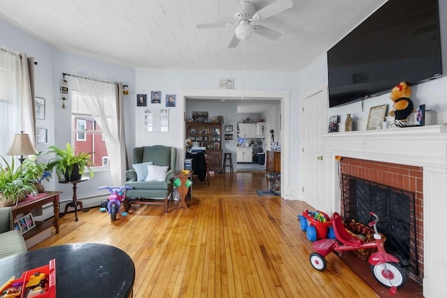 living area with a baseboard heating unit, wood-type flooring, a fireplace, and a ceiling fan