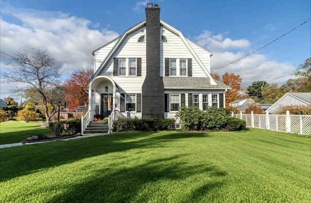 view of front of house with a gambrel roof, a chimney, roof with shingles, fence, and a front yard