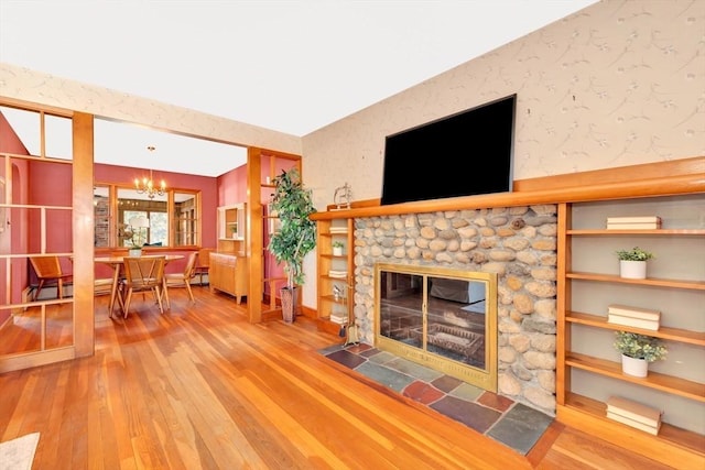 unfurnished living room featuring a notable chandelier, a stone fireplace, wood finished floors, and wallpapered walls