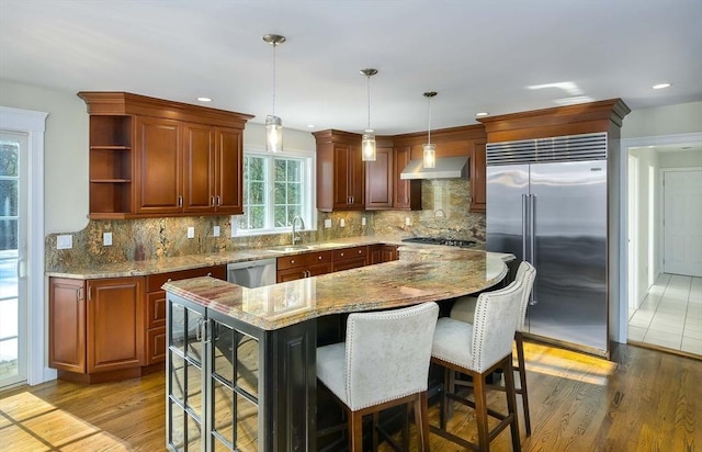 kitchen featuring hanging light fixtures, stainless steel appliances, light stone countertops, a kitchen island, and wall chimney exhaust hood