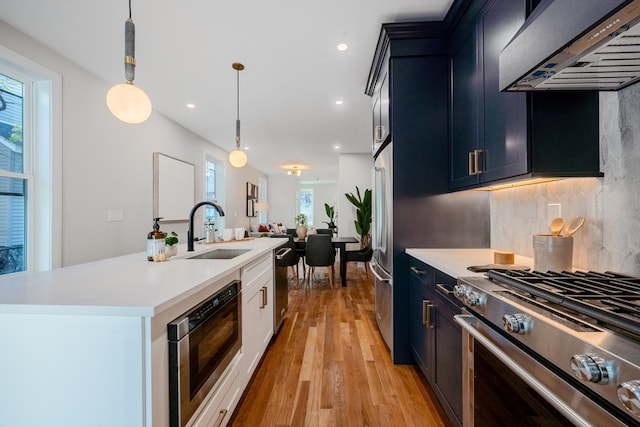 kitchen featuring wall chimney exhaust hood, stainless steel appliances, sink, a center island with sink, and hanging light fixtures