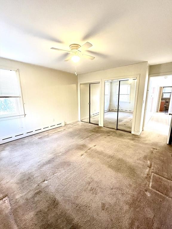 unfurnished bedroom with two closets, ceiling fan, a baseboard radiator, and multiple windows