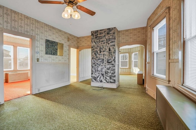 carpeted empty room featuring a wainscoted wall, plenty of natural light, arched walkways, radiator, and wallpapered walls