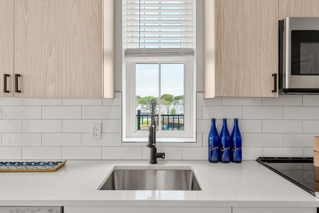 kitchen with light brown cabinetry, a sink, stainless steel microwave, decorative backsplash, and light countertops