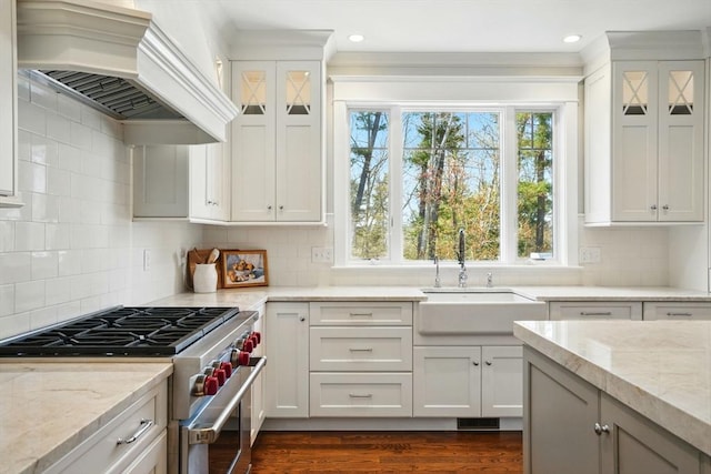 kitchen featuring glass insert cabinets, premium range hood, light stone counters, a sink, and stainless steel range