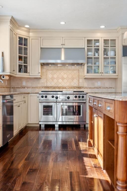 kitchen with dark wood finished floors, glass insert cabinets, cream cabinets, stainless steel appliances, and under cabinet range hood