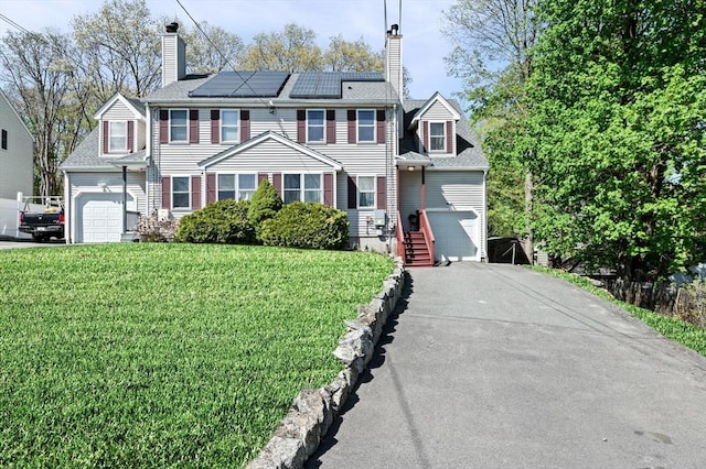colonial-style house featuring a garage, a front lawn, aphalt driveway, a chimney, and roof mounted solar panels