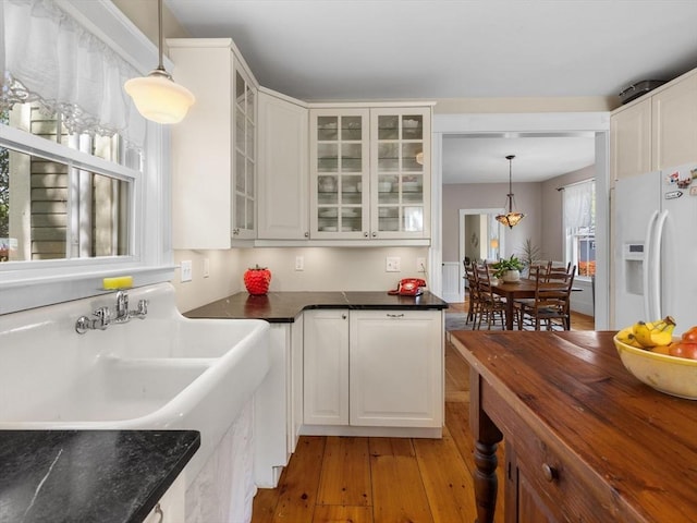 kitchen with dark countertops, hanging light fixtures, light wood finished floors, glass insert cabinets, and white fridge with ice dispenser