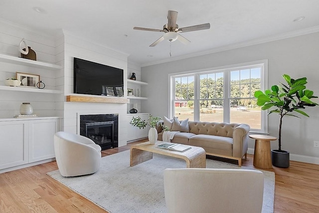 living room with ornamental molding, built in shelves, a large fireplace, ceiling fan, and light hardwood / wood-style floors