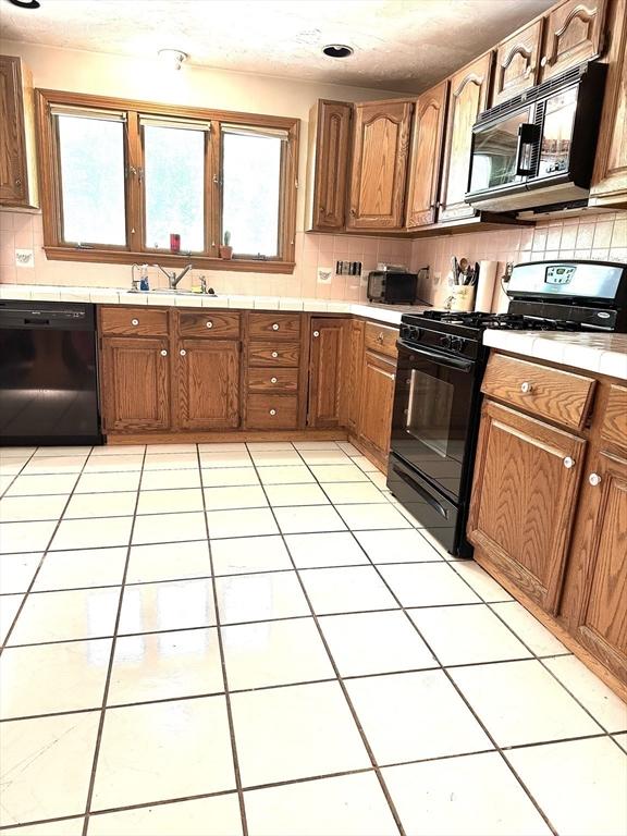 117 Metropolitan Ave Unit 117, Ashland, MA 01721 - kitchen with black appliances, tile counters, tasteful backsplash, brown cabinetry, and light tile patterned flooring