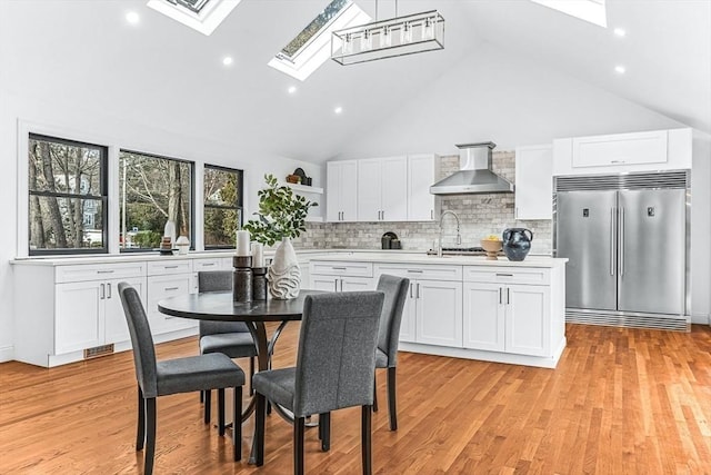 kitchen with a skylight, white cabinetry, light countertops, wall chimney range hood, and stainless steel built in refrigerator