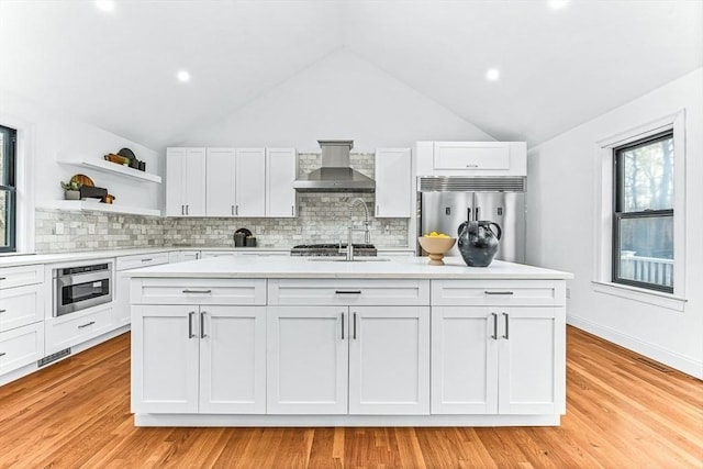 kitchen with white cabinets, appliances with stainless steel finishes, a kitchen island with sink, light countertops, and wall chimney range hood