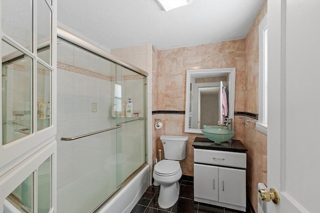 bathroom featuring tile patterned flooring, toilet, bath / shower combo with glass door, vanity, and tile walls