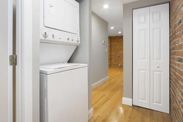 clothes washing area with laundry area, stacked washer and dryer, light wood-type flooring, brick wall, and baseboards