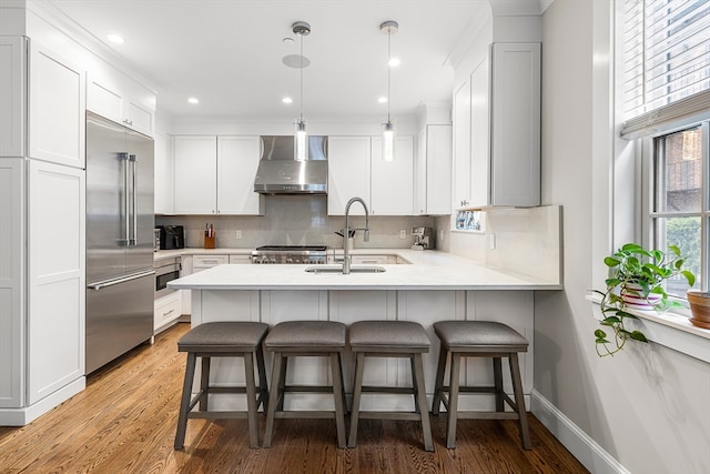 kitchen with wall chimney range hood, sink, stainless steel appliances, hanging light fixtures, and white cabinetry