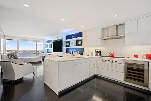 kitchen with wall chimney range hood, wine cooler, kitchen peninsula, white cooktop, and white cabinets