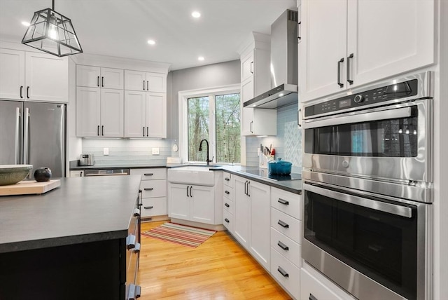 kitchen featuring wall chimney exhaust hood, light wood-type flooring, dark countertops, a sink, and stainless steel appliances