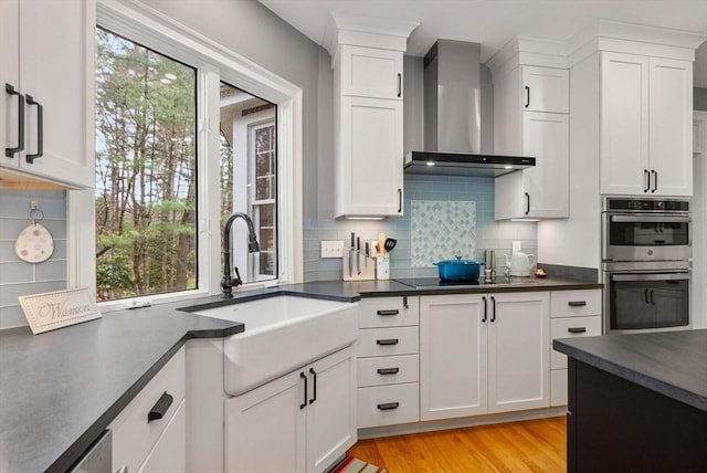 kitchen with dark countertops, wall chimney exhaust hood, black electric cooktop, double oven, and a sink