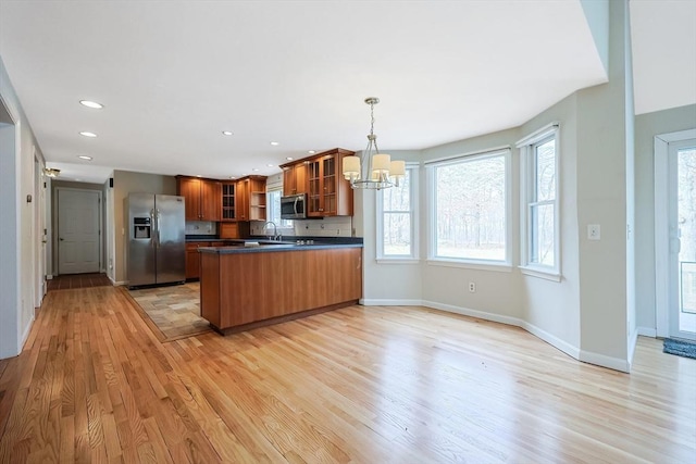 kitchen with dark countertops, appliances with stainless steel finishes, a peninsula, brown cabinetry, and glass insert cabinets