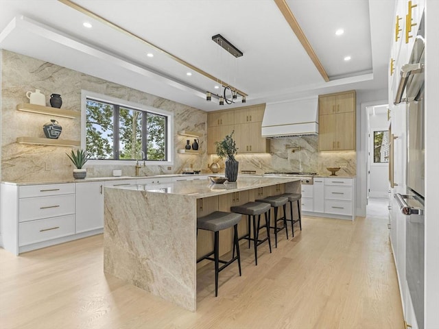 kitchen featuring light stone countertops, a spacious island, white cabinetry, premium range hood, and light hardwood / wood-style flooring