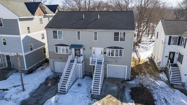 snow covered back of property featuring a garage, roof with shingles, and stairway