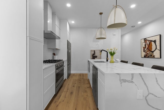 kitchen featuring wall chimney range hood, sink, decorative light fixtures, white cabinetry, and appliances with stainless steel finishes