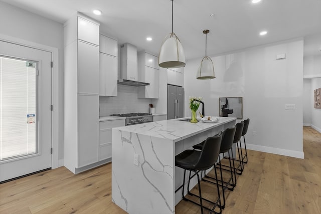 kitchen featuring wall chimney range hood, a center island with sink, pendant lighting, light wood-type flooring, and white cabinets