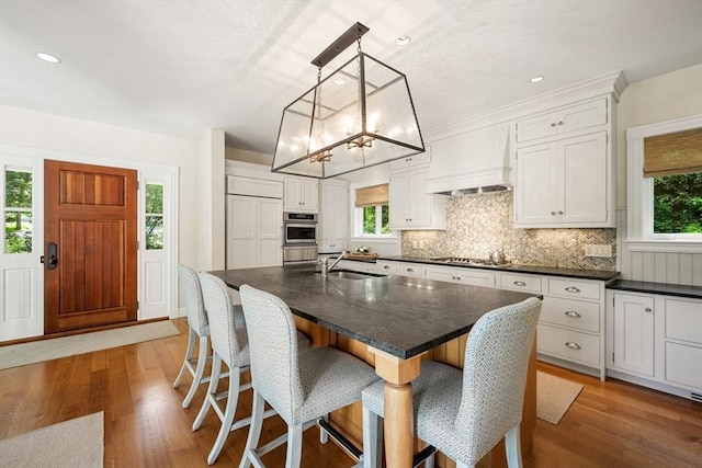 kitchen featuring a center island with sink, dark countertops, custom range hood, hanging light fixtures, and white cabinetry