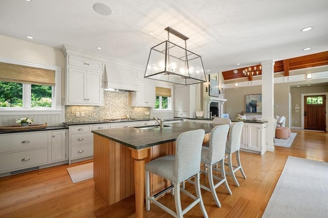 kitchen featuring light wood finished floors, hanging light fixtures, white cabinets, a sink, and wall chimney range hood