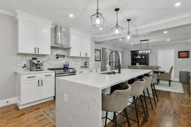 kitchen with hanging light fixtures, wall chimney range hood, a kitchen island with sink, and white cabinets