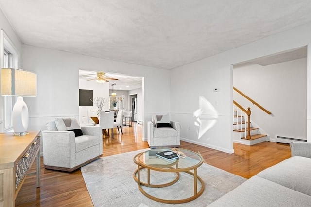 living area with a baseboard heating unit, baseboards, ceiling fan, stairway, and wood finished floors