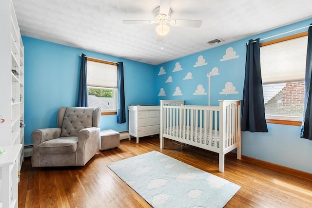 bedroom featuring wood finished floors, a nursery area, baseboard heating, and a ceiling fan