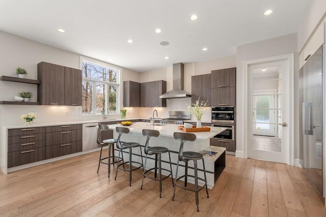 kitchen featuring a breakfast bar area, open shelves, light countertops, appliances with stainless steel finishes, and wall chimney exhaust hood